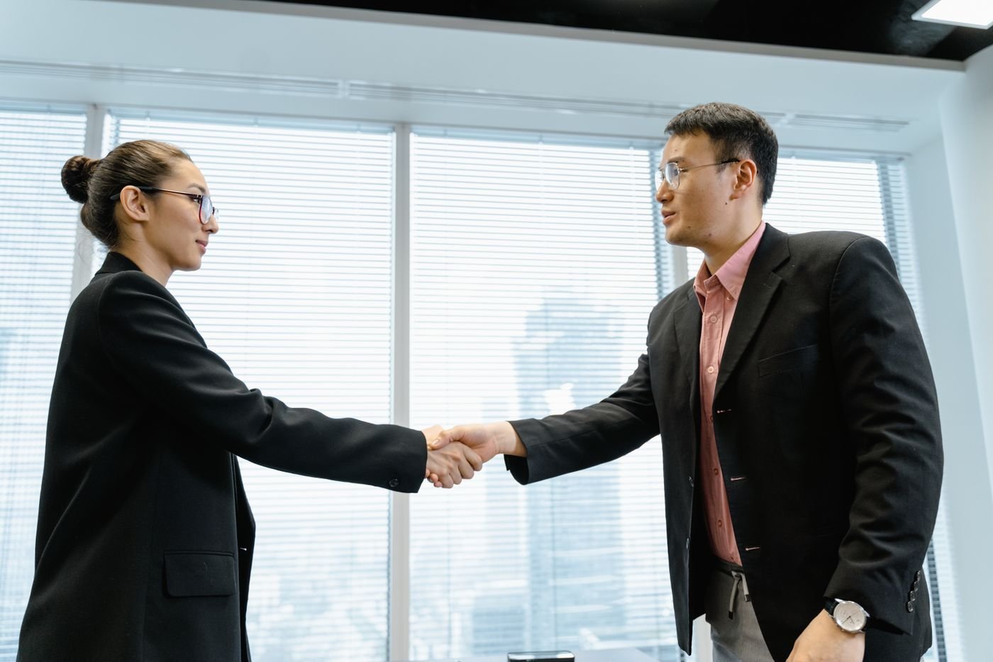 Two business professionals shake hands beside glass office windows.