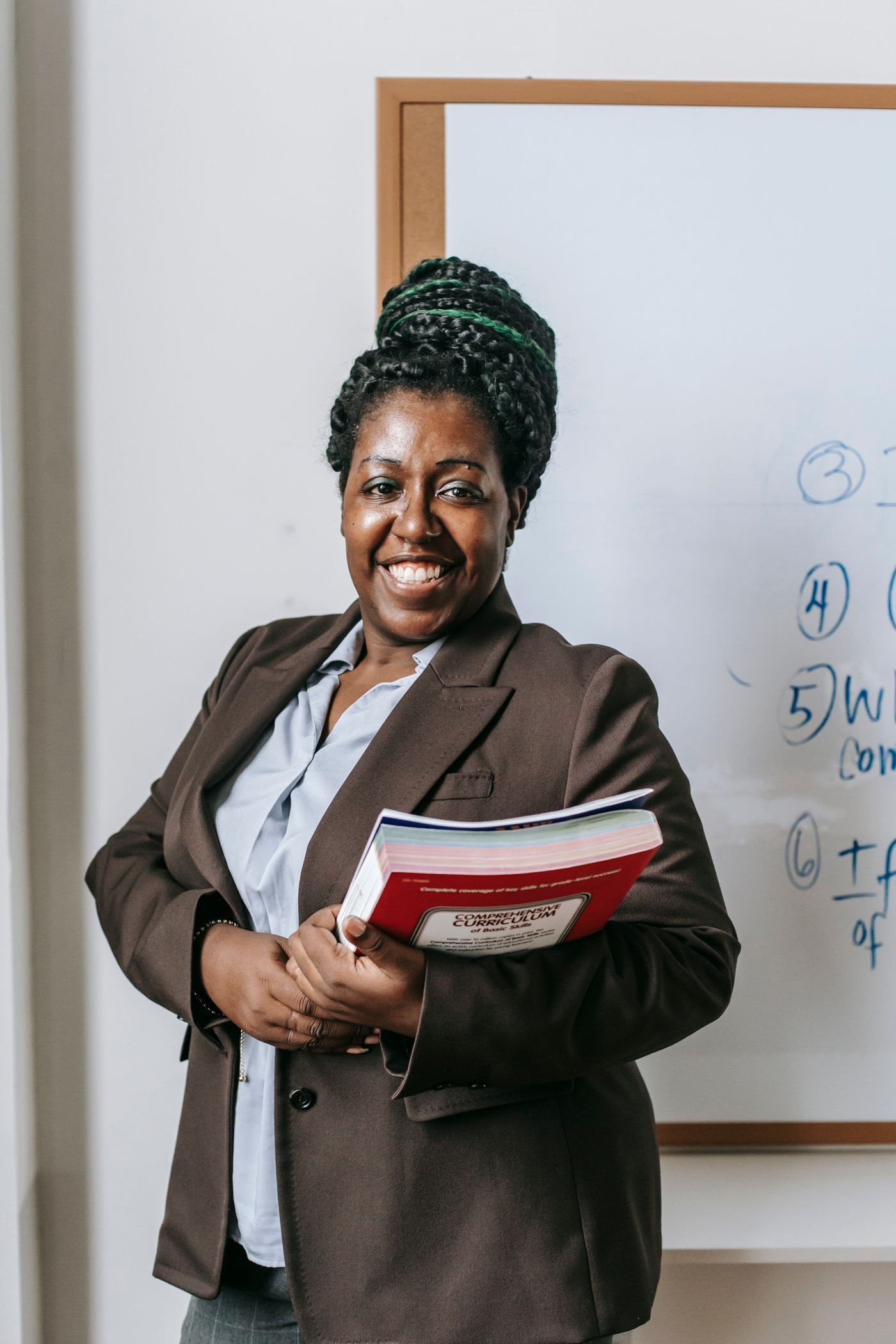 A smiling teacher holds workbooks beside a classroom whiteboard.