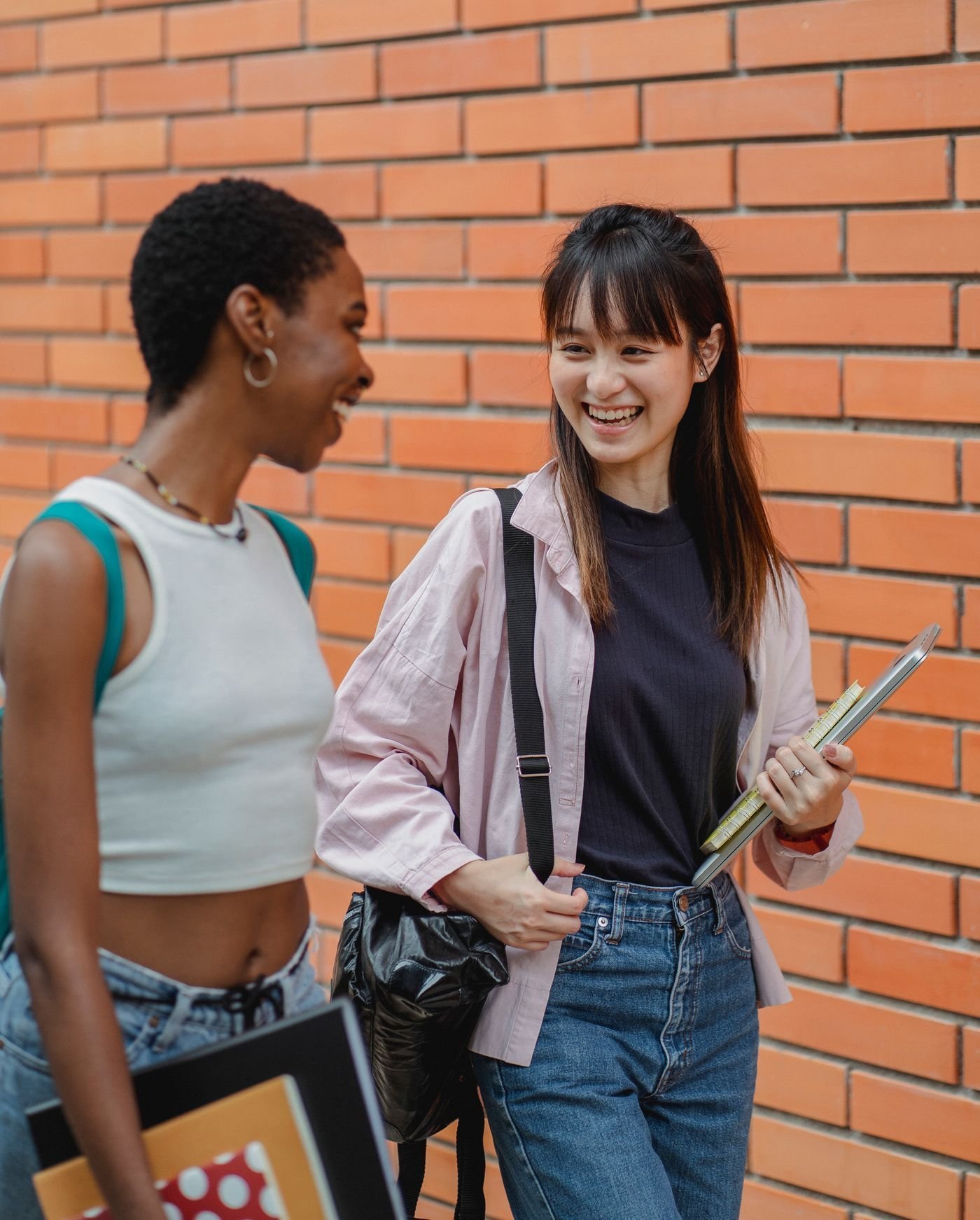 Two smiling students chat beside a brick campus wall.