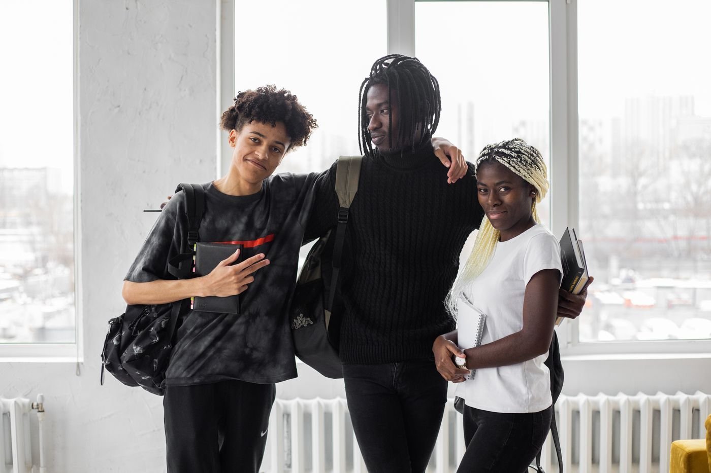 Three smiling students with backpacks stand together near a bright window.